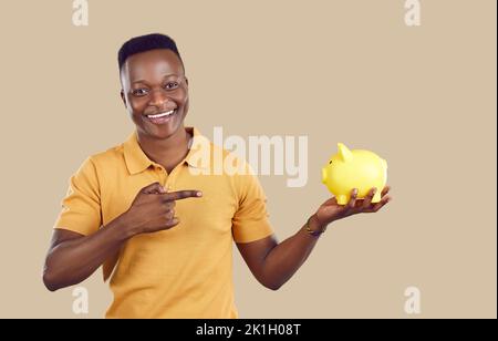 Junger afroamerikanischer Mann zeigt mit dem Finger und hält Sparschwein für Geld auf beigem Hintergrund Stockfoto