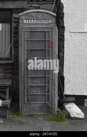 Ein stillgefachter Telefonkasten außerhalb des Kirkstone Pass Inn, Ambleside, Lake District, Großbritannien. Grau, mit roten Farbblitzen und grünem Unkraut. Stockfoto