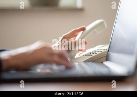 Nahaufnahme der geschäftigen kaukasischen Geschäftsfrau, die auf einem Laptop-Computer tippt, während sie gleichzeitig einen Anruf mit dem klassischen weißen Festnetztelefon tätigen. Stockfoto