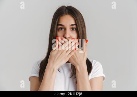 Porträt einer jungen schockierten beiläufigen weißen Frau in T-Shirt mit langen Brünette Haare stehen über grauen Wand Hintergrund bedeckt Mund Stockfoto