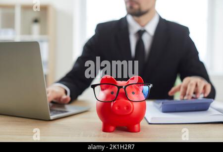 Nahaufnahme des roten Sparschweins in Form von Schweinen steht auf dem Tisch gegen den männlichen Finanzier. Stockfoto