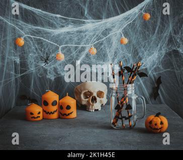 Halloween Tischeinstellung für Getränke. Ein Glas mit Trinkhalmen, Kerzen und einem Totenkopf auf dunklem Grund. Halloween-Dekorationen. Stockfoto