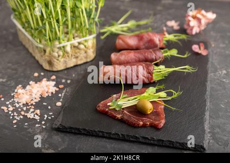 Scheiben geräuchertes gesalzenes Fleisch mit grünem Erbsen-Microgreen auf schwarzem Betongrund. Seitenansicht, Nahaufnahme, selektiver Fokus. Stockfoto