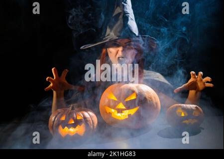 Eine ominöse Hexe im Hut zaubert über eine Jack-o-Laterne. Traditionelle halloween-Figuren. Mystischer Nebel kriecht über Kürbisse mit geschnitzten schrecklich Stockfoto