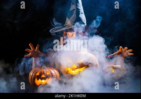 Eine ominöse Hexe im Hut zaubert über eine Jack-o-Laterne. Traditionelle halloween-Figuren. Mystischer Nebel kriecht über Kürbisse mit geschnitzten schrecklich Stockfoto