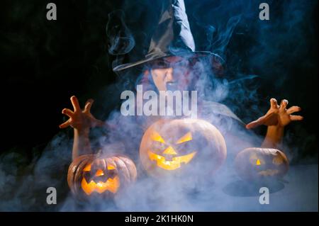 Eine ominöse Hexe im Hut zaubert über eine Jack-o-Laterne. Traditionelle halloween-Figuren. Mystischer Nebel kriecht über Kürbisse mit geschnitzten schrecklich Stockfoto