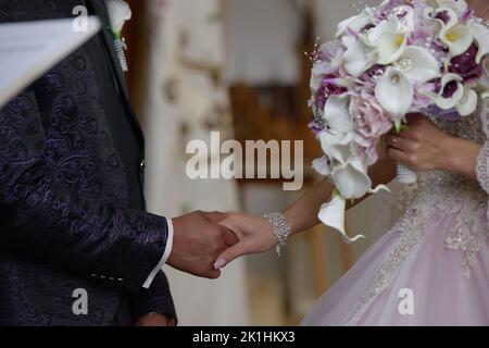 Intimer Moment zwischen einem jungen Paar am Hochzeitstag, Braut in einem lilafarbenen Kleid und mit einem übergroßen Blumenstrauß, während sie die Hände hält Stockfoto