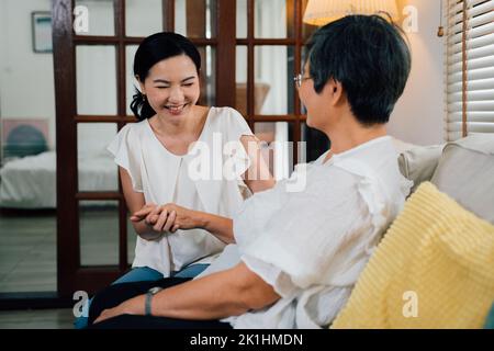 Fröhliche Erwachsene asiatische Tochter lächelt und hält die Hand der alten Mutter, während sie am Wochenende zu Hause auf dem Sofa im Wohnzimmer sitzt Stockfoto