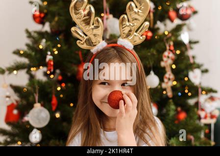 Ein kleines Mädchen im Geweih eines Weihnachtshirsches hält ihr einen Ball an die Nase. Frohes neues Jahr und weihnachten Stockfoto