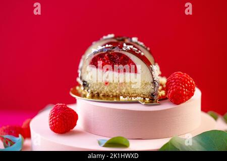 Beerendessert. Runder Kuchen mit Himbeerfüllung geschnitten und Himbeeren auf einem rosafarbenen Podium auf rotem Hintergrund.zarter, luftiger Kuchensouffle.Choux-Gebäck. Stockfoto