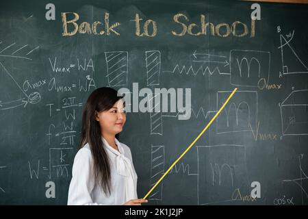 Zurück zum Schulkonzept. Asiatische Lehrerin lächelnd mit Holzstab zeigt auf Tafel in der Schule im Klassenzimmer, glücklich schöne junge Frau Stan Stockfoto