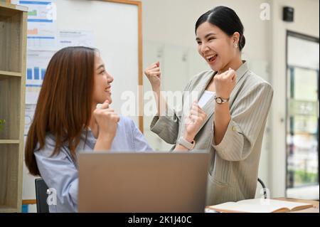 Überfreudige, tausendjährige asiatische Geschäftsfrauen feiern Geschäftserfolg, erhalten ein Geschäft viel und brüllen gemeinsam im Büro vor Freude. Stockfoto