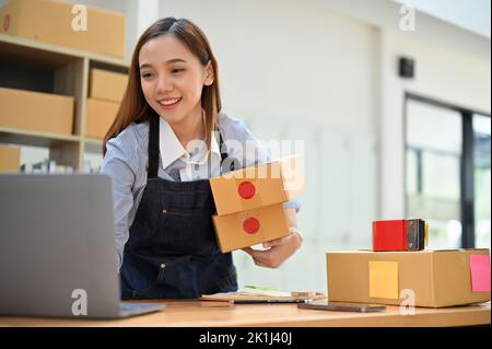 Erfolgreiche Millennial asiatische weibliche Online-Shop-Besitzer oder Kleinunternehmen Startup Überprüfung der Online-Bestellungen des Kunden auf Laptop während der Vorbereitung Versand pa Stockfoto