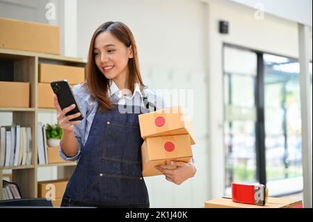 Wunderschöne junge asiatische weibliche Online-Shop-Besitzer oder Unternehmen Startup mit ihrem Smartphone, während sie ihre Versandpakete in ihrer Stock Office. E-comm Stockfoto