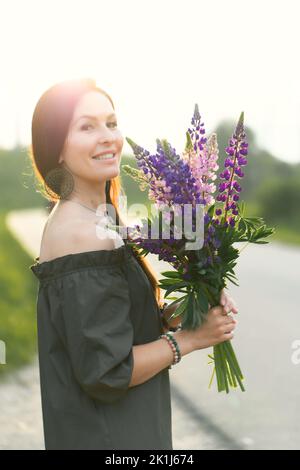 Attraktive Frau auf der Straße in einem Sommerfeld unter Lupinus Blumen. Vertikales Foto. Stockfoto