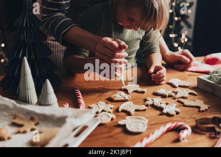 Weihnachten und Neujahr Lebensmittelzubereitung. Weihnachten Lebkuchen Kochen und dekorieren frisch gebackene Kekse mit Glasur und Mastix. Mama hilft niedlichen kleinen Tochter Cookie auf Holz chaotisch Tisch zu schmücken Stockfoto