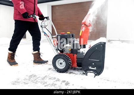 Detailaufnahme eines tragbaren roten Schneefräsers, angetrieben von Benzin in Aktion. Älterer älterer Mann im Freien vor dem Haus, der nach dem Schneesturm Schneefräse benutzt, Schneefall zum Abräumen verwendet und Schnee entfernt Stockfoto