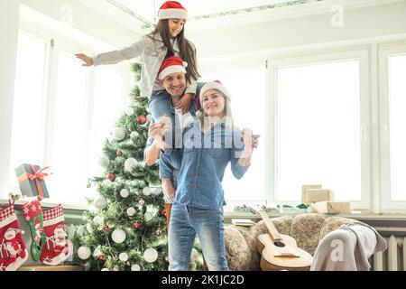 Familie schmücken einen Weihnachtsbaum. Junger Mann mit seiner Tochter auf seinen Schultern hilft ihr, den Weihnachtsbaum schmücken. Stockfoto