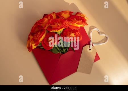 Schöne rote Rosen Blumen in postalischem roten Umschlag auf rotem Hintergrund, beige Papiernote und Seil in Herzform Kopieplatz für Text, Frühling, Grußkarte für Urlaub. Blumenlieferung. Zarte rote gelbe Rosen. Monochrome Minimal trendige Komposition. Romantische Blumen Stockfoto