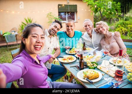 Glückliche asiatische Frau, die ein Selfie-Foto mit Smartphone mit verschiedenen erwachsenen Freunden posiert für das Bild. Eine multiethnische Gruppe von Männern und Frauen Stockfoto