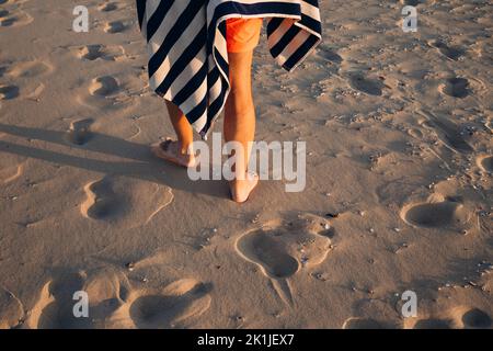 Der junge Kaukasusmann in roten Badeshorts mit gestreiften Handtüchern spaziert entlang des sandigen Sommerstrands. Männliche Beine in Nahaufnahme auf Sand bei Sonnenuntergang. Stockfoto