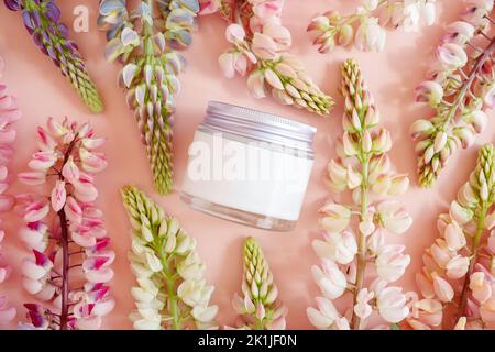 Feuchtigkeitsspendende Creme im Glas mit Schraubverschluss aus Metall und rosa Lupinenblüten viel auf rosa Hintergrund. Modell, Vorderansicht von oben. Kosmetische Feuchtigkeitscreme bo Stockfoto