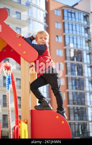 Männliches Kind, das auf der Rutsche steht, die Kamera anschaut und sich im Spielbereich mit dem Kopf an der Hand lehnt. Netter Junge klettert auf Spielplatzstruktur, posiert an sonnigen Tag. Moderne Wohngebäude im Hintergrund. Stockfoto