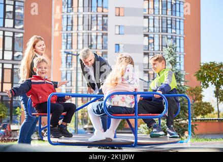 Glückliche Familie - Vater, Mutter und Kinder, die Spaß zusammen im modernen Innenhof der Stadt Wohnhochhäuser haben. Lächelnde Eltern, die mit Kindern spielen und auf dem Spielplatz schaukeln. Stockfoto