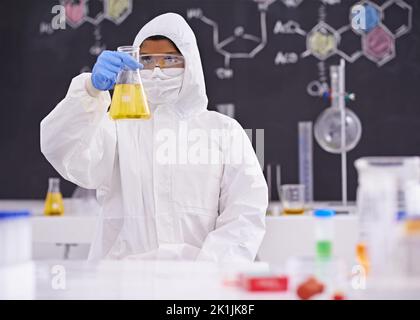 Warten auf die Reaktion. Eine junge Wissenschaftlerin in Schutzkleidung, die in ihrem Labor eine Flasche hochhält. Stockfoto