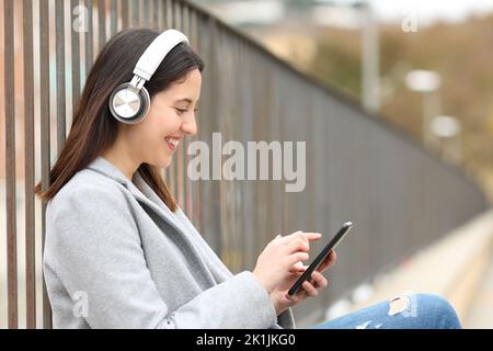 Seitenansicht Porträt einer glücklichen Frau, die mit Kopfhörern Musik hört und das Telefon auf der Straße benutzt Stockfoto