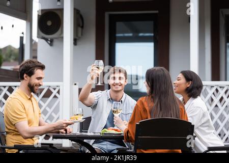 Glücklicher Mann, der ein Glas Wein in der Nähe von interracial Freunden während der bbq-Party auf dem Hinterhof anhebt Stockfoto