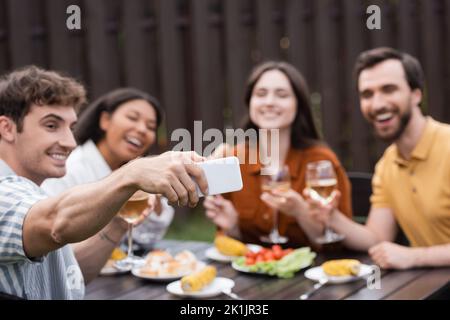 Fröhlicher Mann, der während der grillparty mit verschwommenen multiethnischen Freunden Selfie gemacht hat Stockfoto