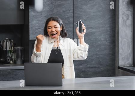 Aufgeregt bi-rassische Frau hält Smartphone und Blick auf Laptop in der Küche Stockfoto