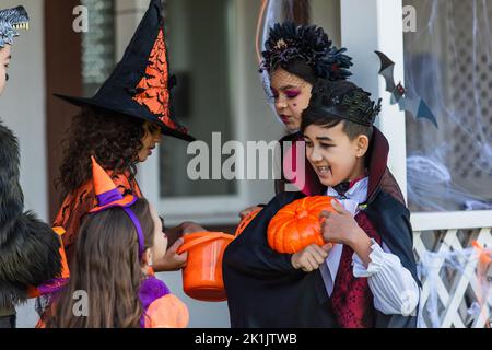 Lächelnder asiatischer Junge mit Make-up, der auf Kürbis in der Nähe von Freunden in halloween-Kostümen im Hinterhof zeigt Stockfoto