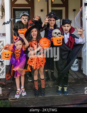 Multiethnische Kinder in halloween-Kostümen halten Eimer und Grimassen vor der Kamera in der Nähe des Hauses Stockfoto