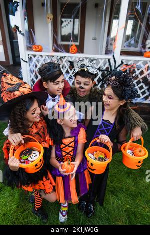 Fröhliche multiethnische Kinder in halloween-Kostümen halten Eimer mit Süßigkeiten und Kürbis im Hinterhof Stockfoto