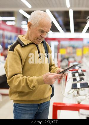 Älterer grauhaariger Rentner, der im Ausstellungsraum des digitalen Warenhauses einen Schalter mit elektronischen Geräten und Tablets untersucht Stockfoto