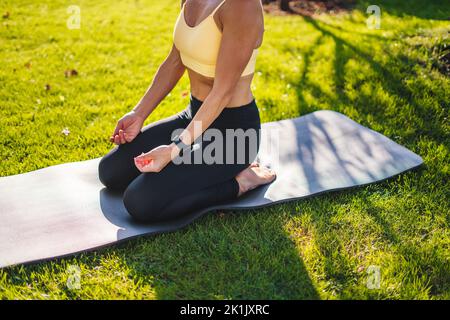 Beschnittene Ansicht einer Frau, die Yoga praktiziert und in einer bequemen Sitzposition auf einer Matte sitzt und im Sommerpark meditiert. Yoga-Übung, Dehnung der Oberschenkel. Körperform Stockfoto