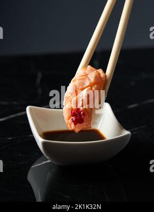 Nahaufnahme mit Bambusstäbchen und Lachs-Nigiri, während Sie Nigiri im Restaurant in Sojasauce tauchen. Vorderansicht. Essen, japanische Küche c Stockfoto