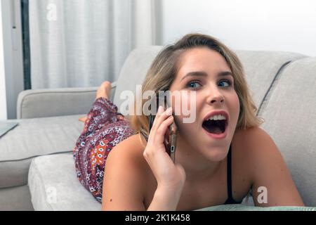 Junge Frau, die auf dem Sofa liegt und mit einem erstaunten Gesichtsausdruck am Telefon spricht Stockfoto