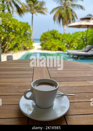 Konzentrieren Sie sich auf eine Tasse Kaffee in einer weißen Tasse mit einem unscharfen tropischen Inselresort auf den Malediven. Stockfoto