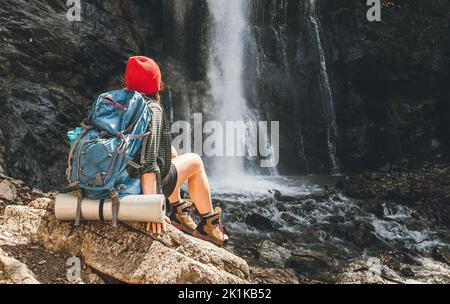 Frau mit einem Rucksack in einem roten Hut in aktiven Trekking-Kleidung in der Nähe des Bergflußwasserfalls sitzen und genießen Sie die plätschernden Natur powe gekleidet Stockfoto