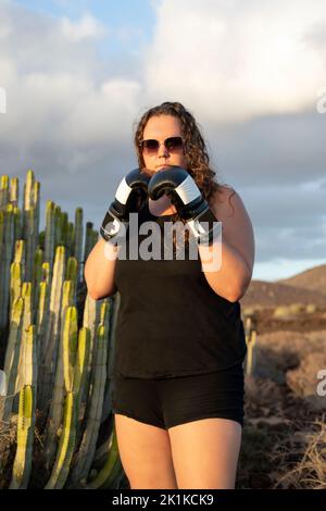 Eine junge, gesunde weiße Frau, die eine Sonnenbrille, schwarze Sportbekleidung und Boxhandschuhe trägt und Hände und Arme in einer Kampfposition hält Stockfoto