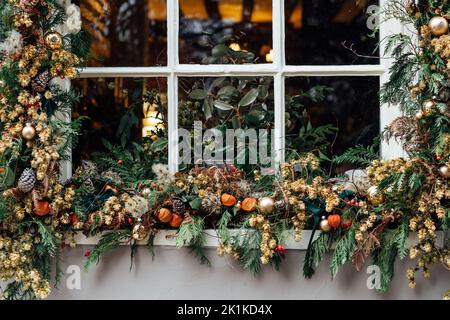 Das Fenster für den Winterurlaub ist mit frischem Grün, roten Beeren, Zapfen und Blumen geschmückt. Außendekoration aus natürlichen Materialien. Weihnachten fe Stockfoto