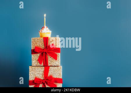 Festlicher Cupcake mit Birning Candle auf dem Turm aus golden glitzernden Geschenkschachteln mit rotem Samtband auf blauem Hintergrund. Feiern Stockfoto