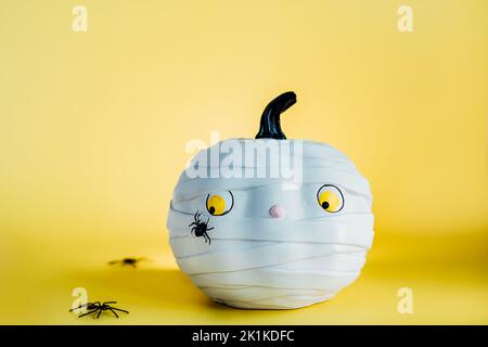 Lustige und niedliche Kürbis-Mumie mit Spinne auf dem gelben Hintergrund. Weißer Kürbis mit beängstigtem und besorgtem Look. Angst vor Insekten. Halloween-Dekor und b Stockfoto