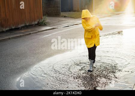 Frau, die nach dem Regen auf der Straße Spaß hat. Lächelnde Frau mit Regengummistiefeln und gelbem Regenmantel, die mit Wasserspritzern und Wasserspritzern in die Pfütze läuft Stockfoto