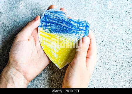 Ansicht von oben auf die Hände, die eine papierförmige ukrainische Flagge in Herzform halten Stockfoto