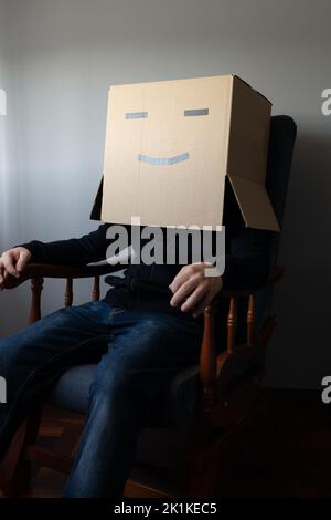 Lächelnder Mann mit einem Pappkarton auf dem Kopf, der auf einem Stuhl sitzt Stockfoto