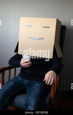 Lächelnder Mann mit einem Pappkarton auf dem Kopf, der mit seinem Smartphone auf einem Stuhl sitzt Stockfoto